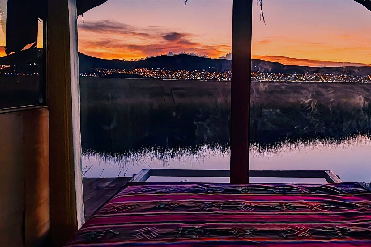 Una habitación con una cama, una ventana con vistas a un paisaje urbano al atardecer y un adorno en la pared.