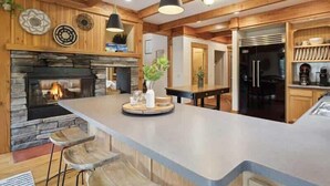 Una cocina moderna con una isla grande, vigas de madera, una chimenea de piedra y un refrigerador empotrado.