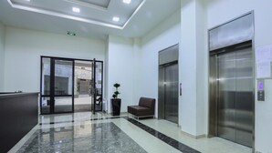 A modern lobby with a marble floor, two elevators, a reception desk, and a seating area.