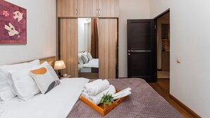 A hotel room with a bed, a bedside table with a lamp, a wooden wardrobe, a mirror, and a door leading to a bathroom.
