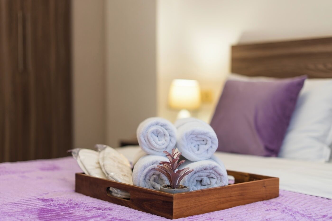 A wooden tray with rolled towels and a potted plant on a bed with a purple blanket.
