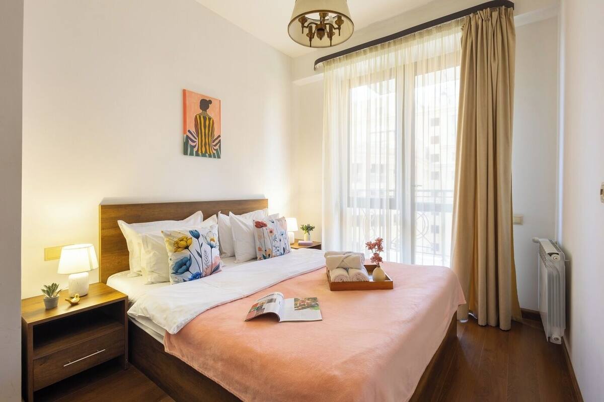 A hotel room with a bed, a nightstand, a lamp, a window with curtains, and a radiator.
