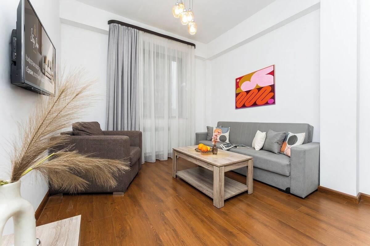 A living room with a grey sofa, a coffee table with fruit, a TV on the wall, and a large window with sheer curtains.
