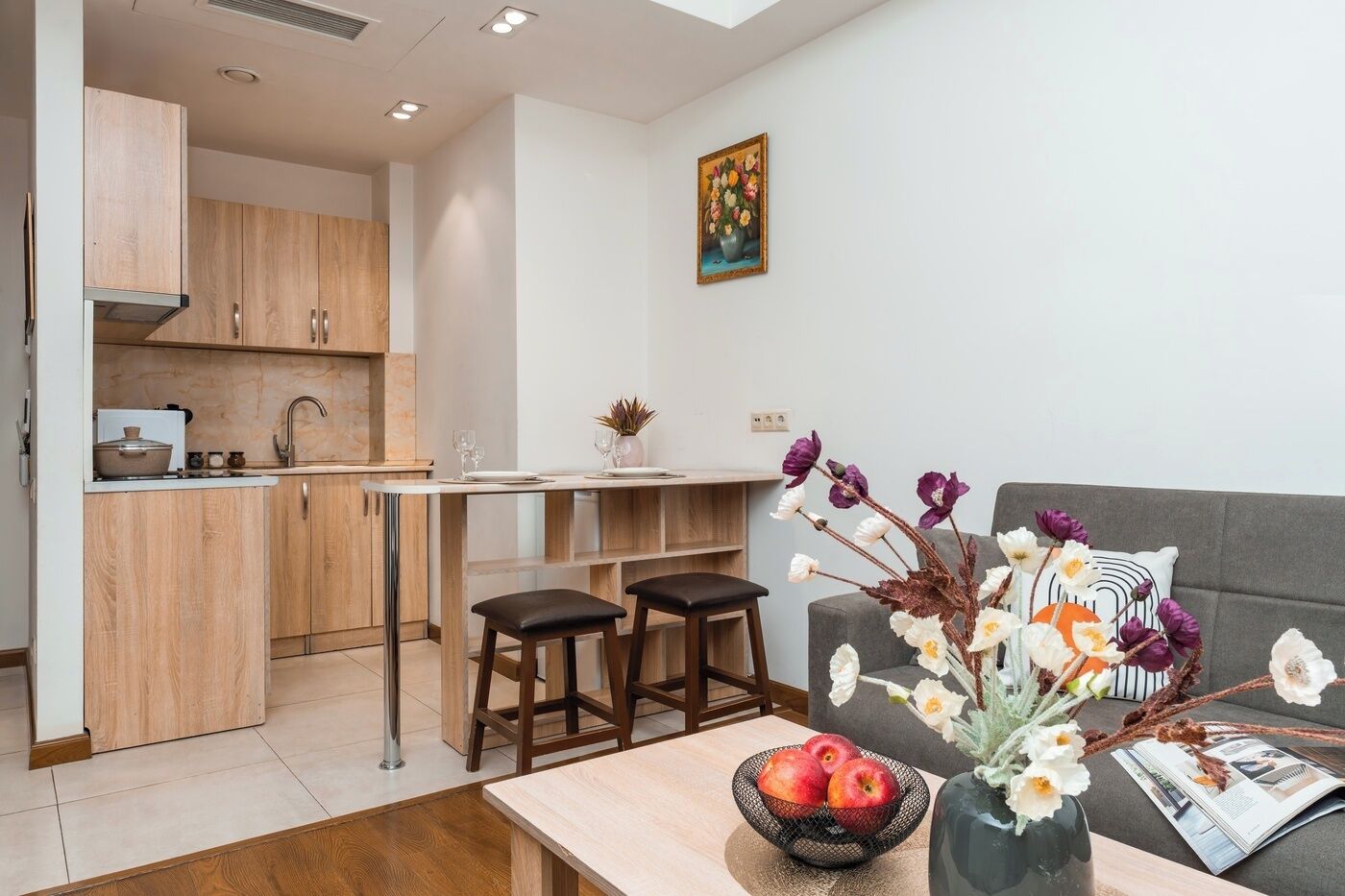 A modern kitchen with wooden cabinets, a dining area with a table and chairs, and a living area with a sofa and decorative flowers.