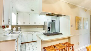 Uma cozinha compacta com armários brancos, geladeira de aço inoxidável, cafeteira e uma pequena ilha com banquetas.