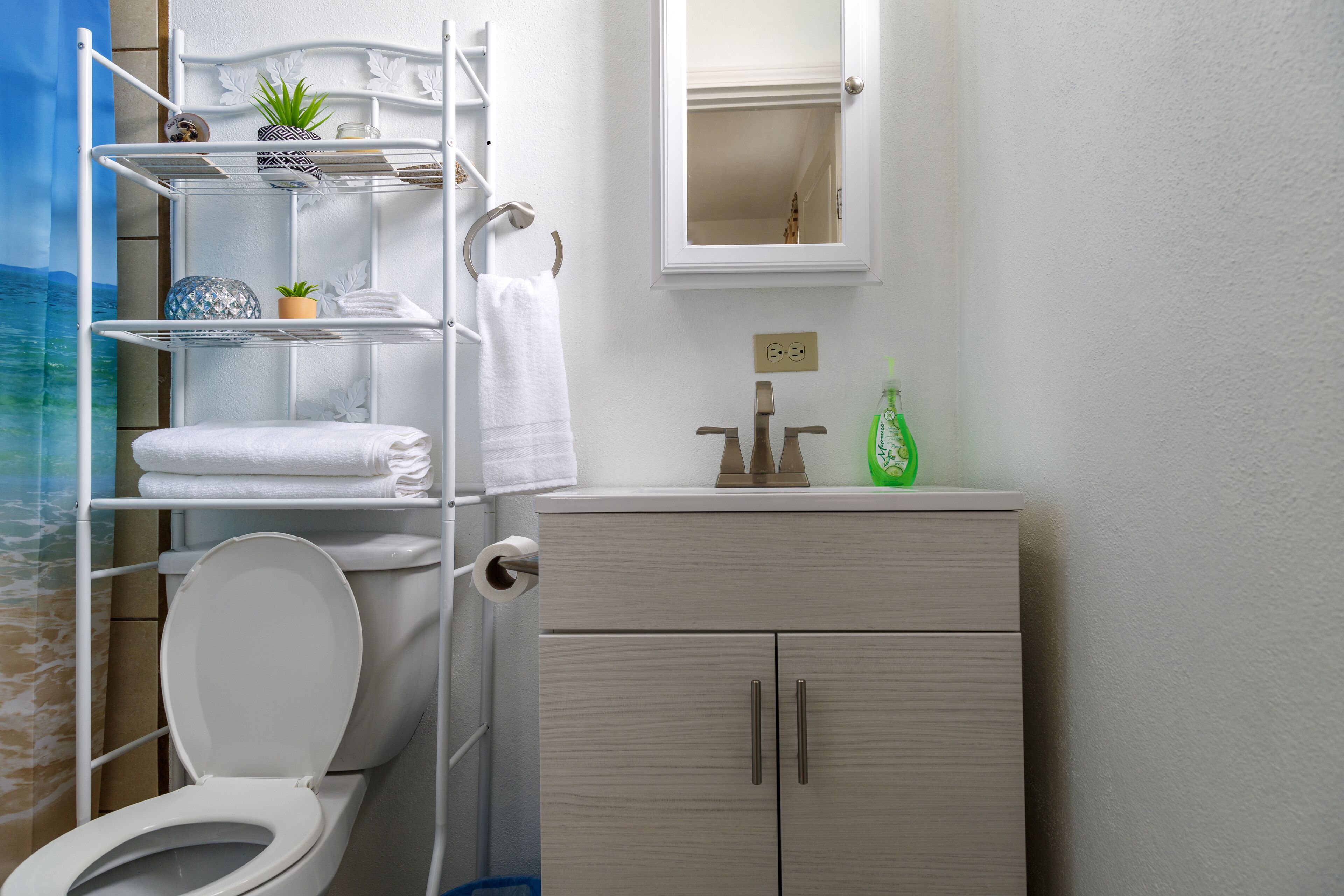 Un baño con inodoro, lavamanos, espejo y repisas con toallas y artículos de aseo.