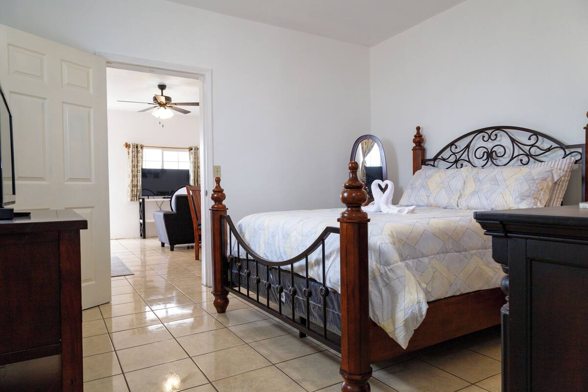 A bedroom with a wooden bed, a ceiling fan, a mirror, and a view of a living area with a TV and a chair.