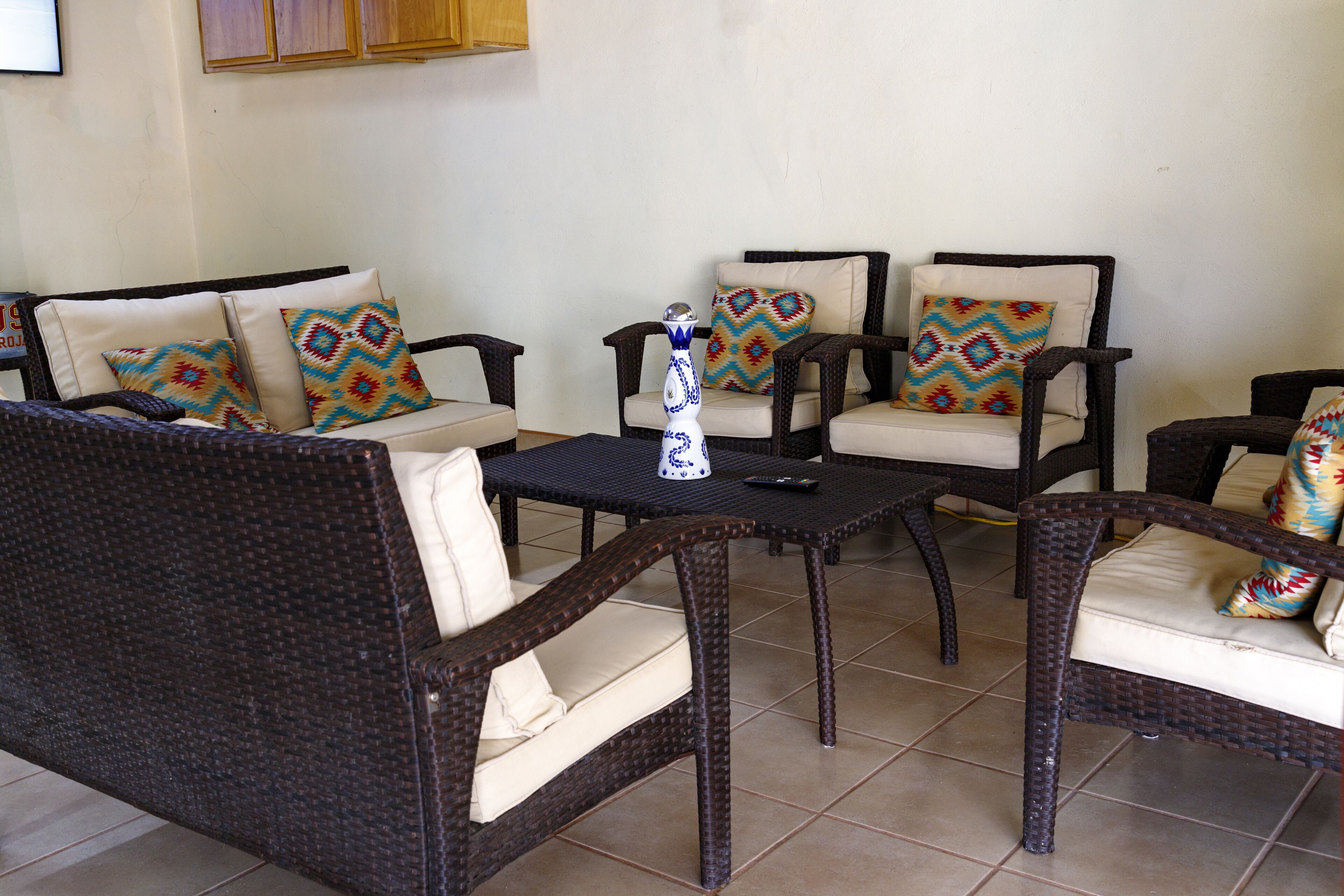 A seating area with wicker furniture, including two sofas and two armchairs, each with patterned cushions. A small table with a vase and remote control is in the center.
