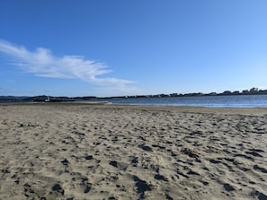A sandy beach with footprints, a clear blue sky, and a distant shoreline with buildings. - Cozy Cottage • Fire Pit • Walk to Bay (Lincoln City)