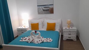 A neatly made bed with a blue bedspread, white pillows, and a swan-shaped towel arrangement. There are two bedside tables with lamps, and a painting of a boat on the wall. - Gennadi Retreat - House 2 (Rhodes)