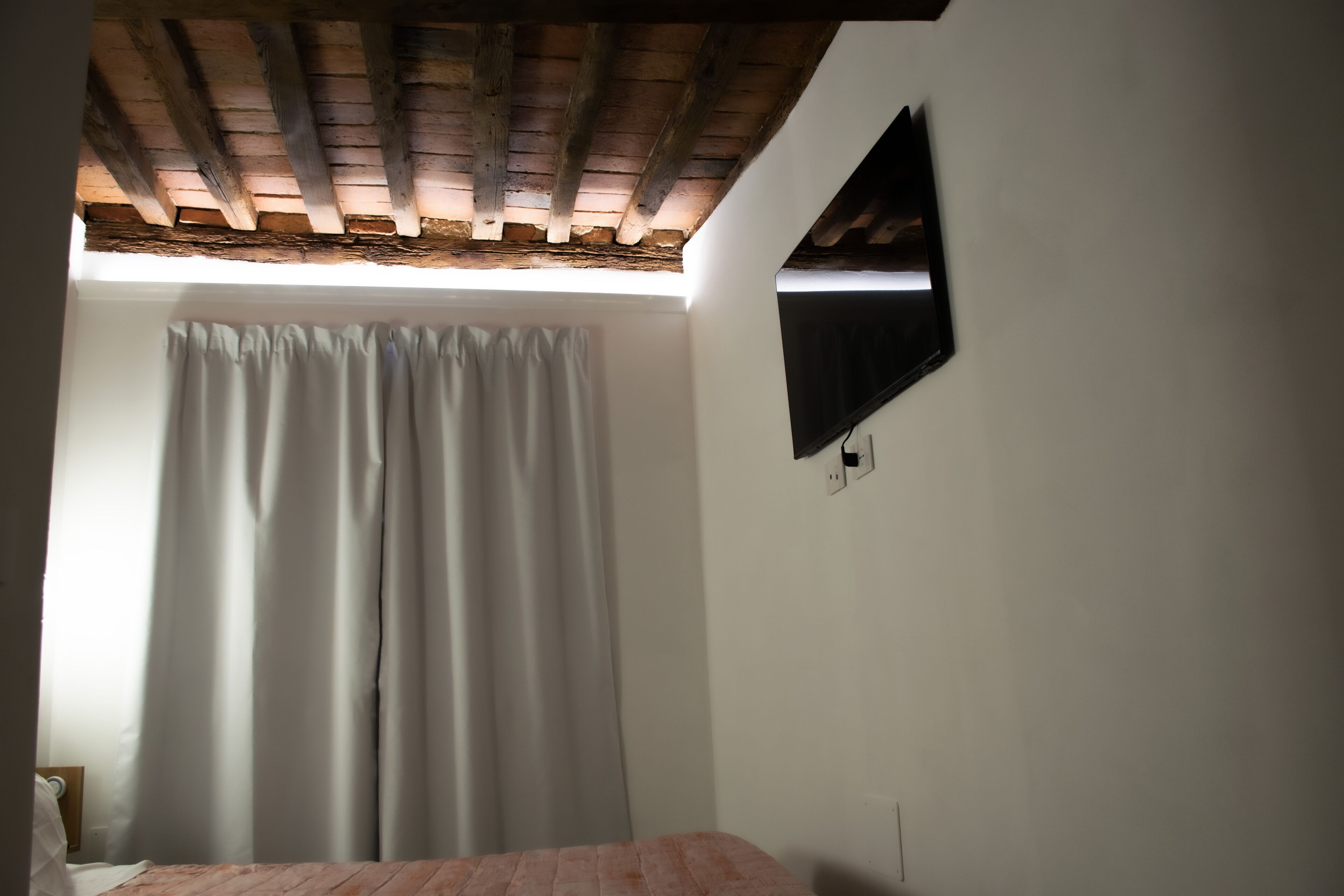 Une chambre avec un plafond en bois, un lit, une fenêtre avec des rideaux blancs et un téléviseur fixé au mur.