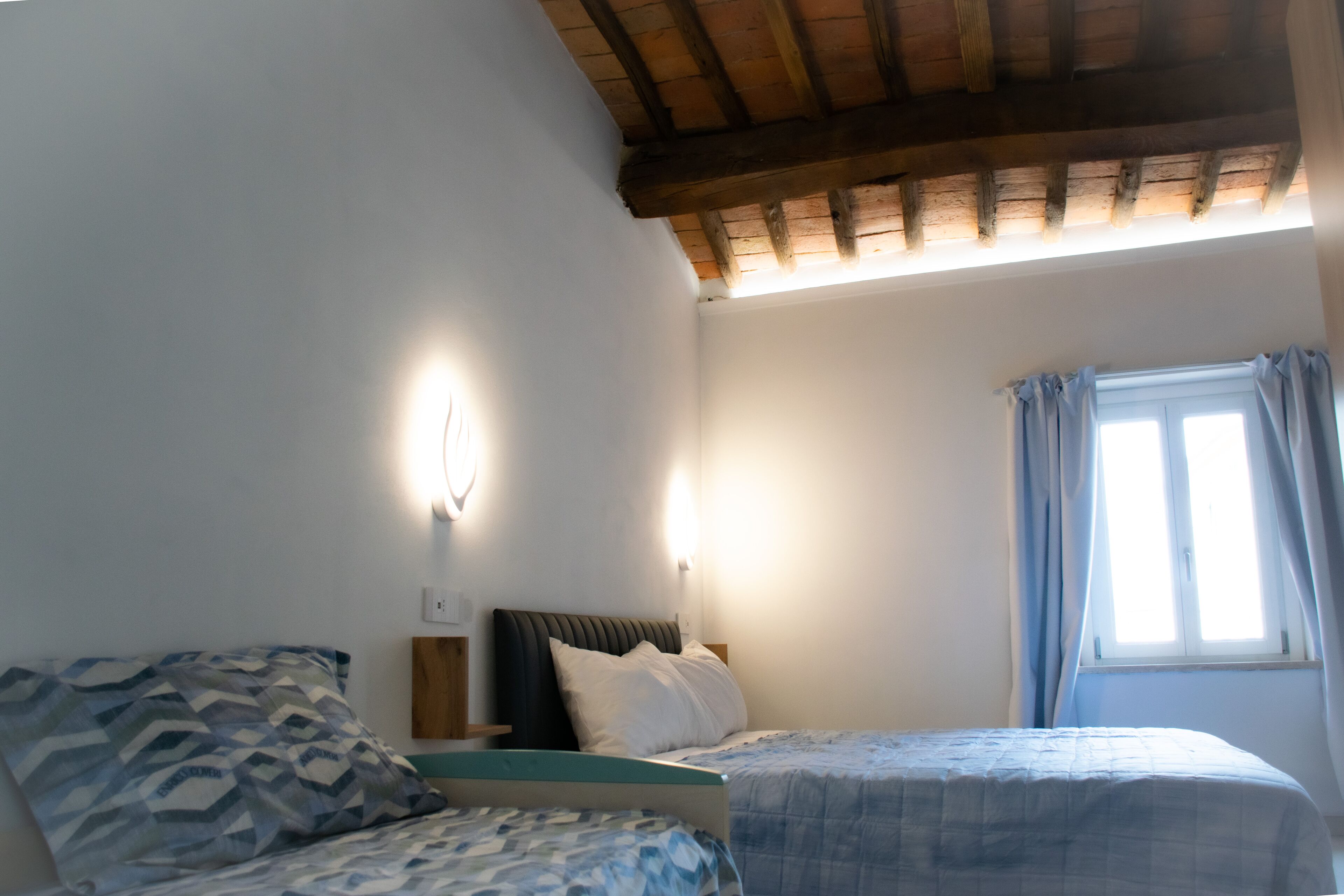 A bedroom with two beds, a wooden ceiling, a window with curtains, and a wall-mounted light.
