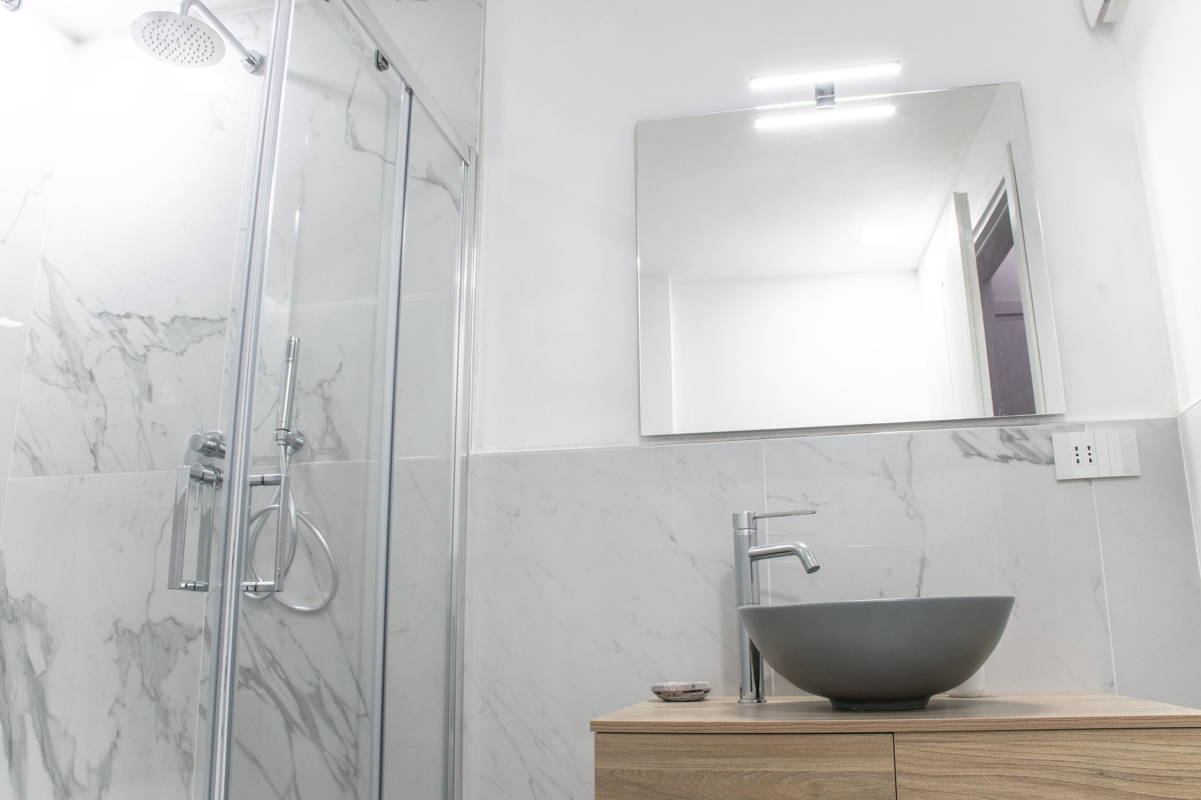 A modern bathroom with a glass shower enclosure, a marble-tiled wall, a wooden vanity, and a large mirror.