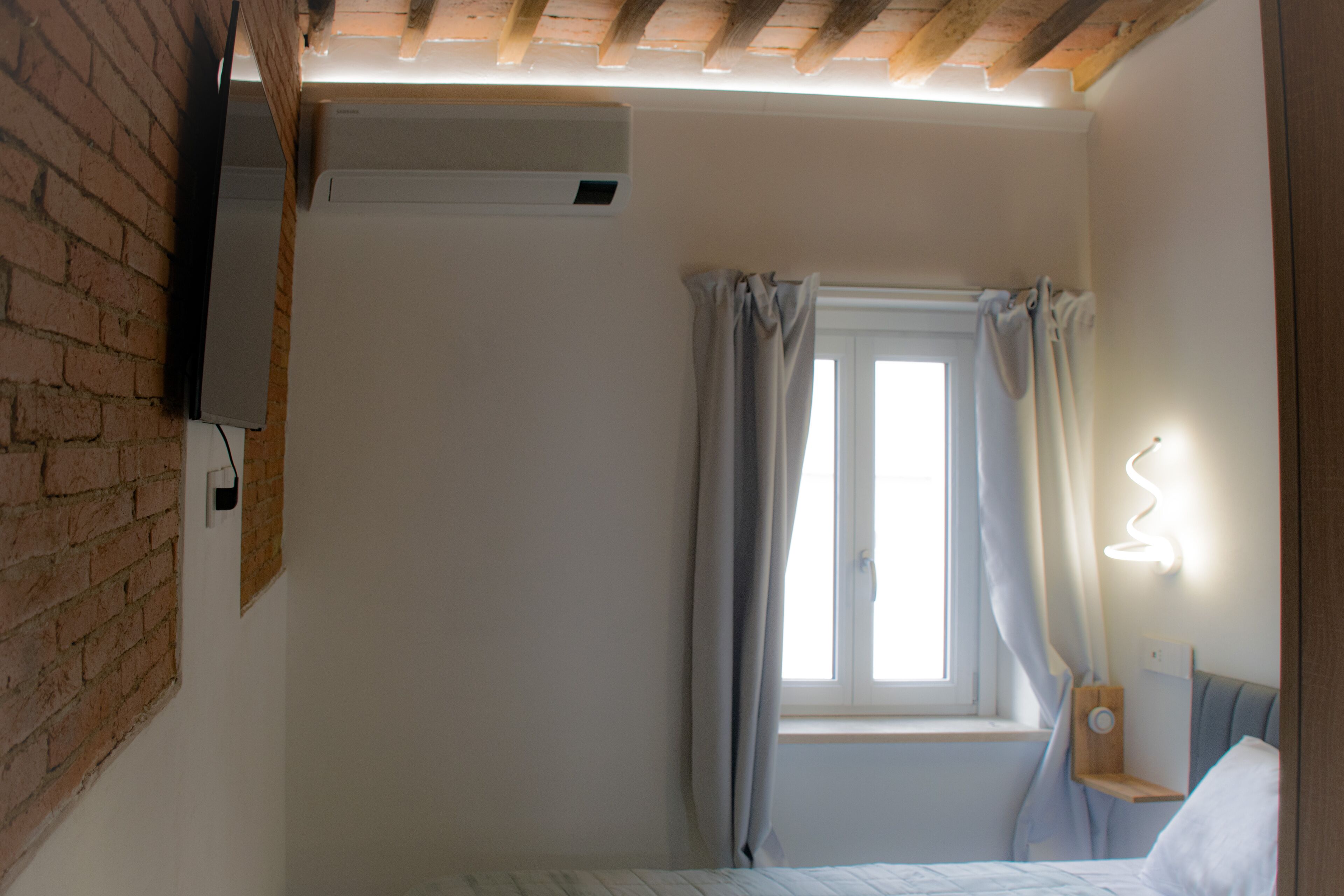 A bedroom with a brick wall, an air conditioning unit, a window with curtains, and a bedside table with a lamp.