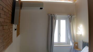 A bedroom with a brick wall, an air conditioning unit, a window with curtains, and a bedside table with a lamp.