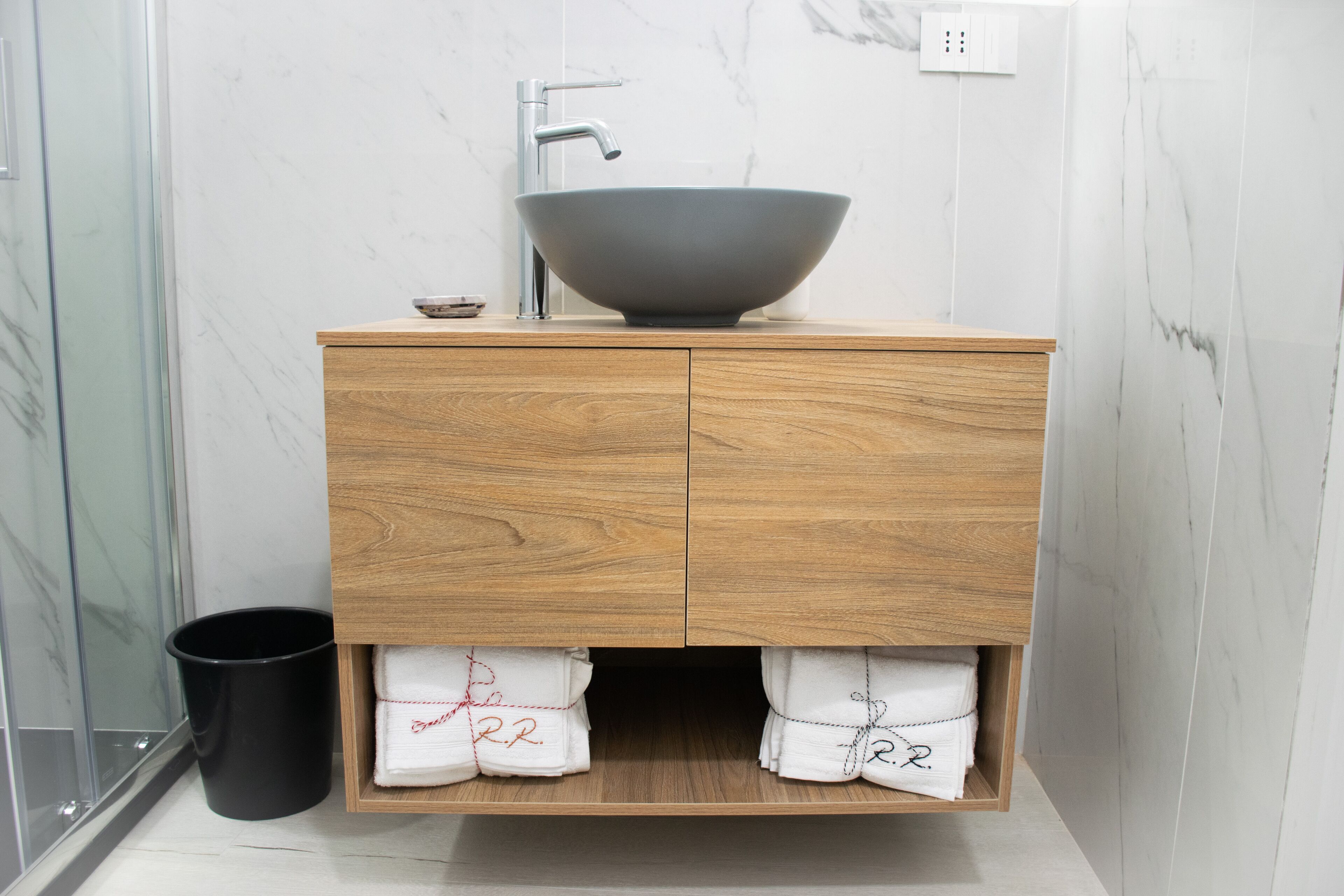 A modern bathroom with a wooden vanity, a large vessel sink, and marble walls.