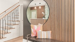 Una escalera con escalones de madera y barandillas negras, un espejo redondo que refleja una habitación con un revestimiento de listones de madera, y dos cojines sobre una mesa oscura.