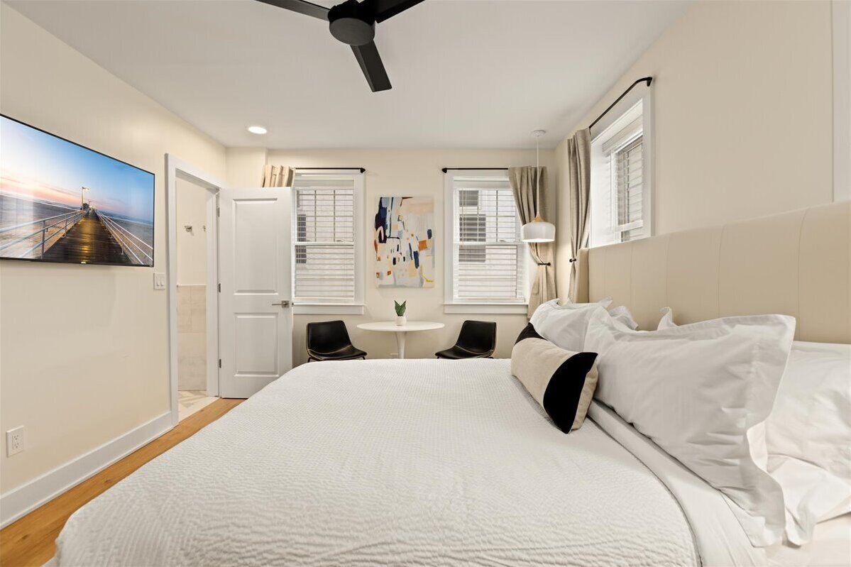 A modern hotel room with a large bed, a ceiling fan, a small table with chairs, and a painting on the wall.