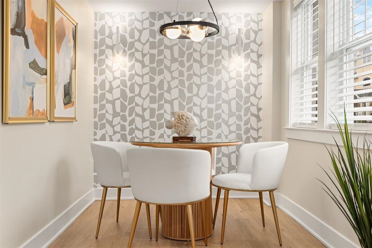 A dining area with a round table, four white chairs, a wooden floor, a patterned wallpaper, and a hanging light fixture.
