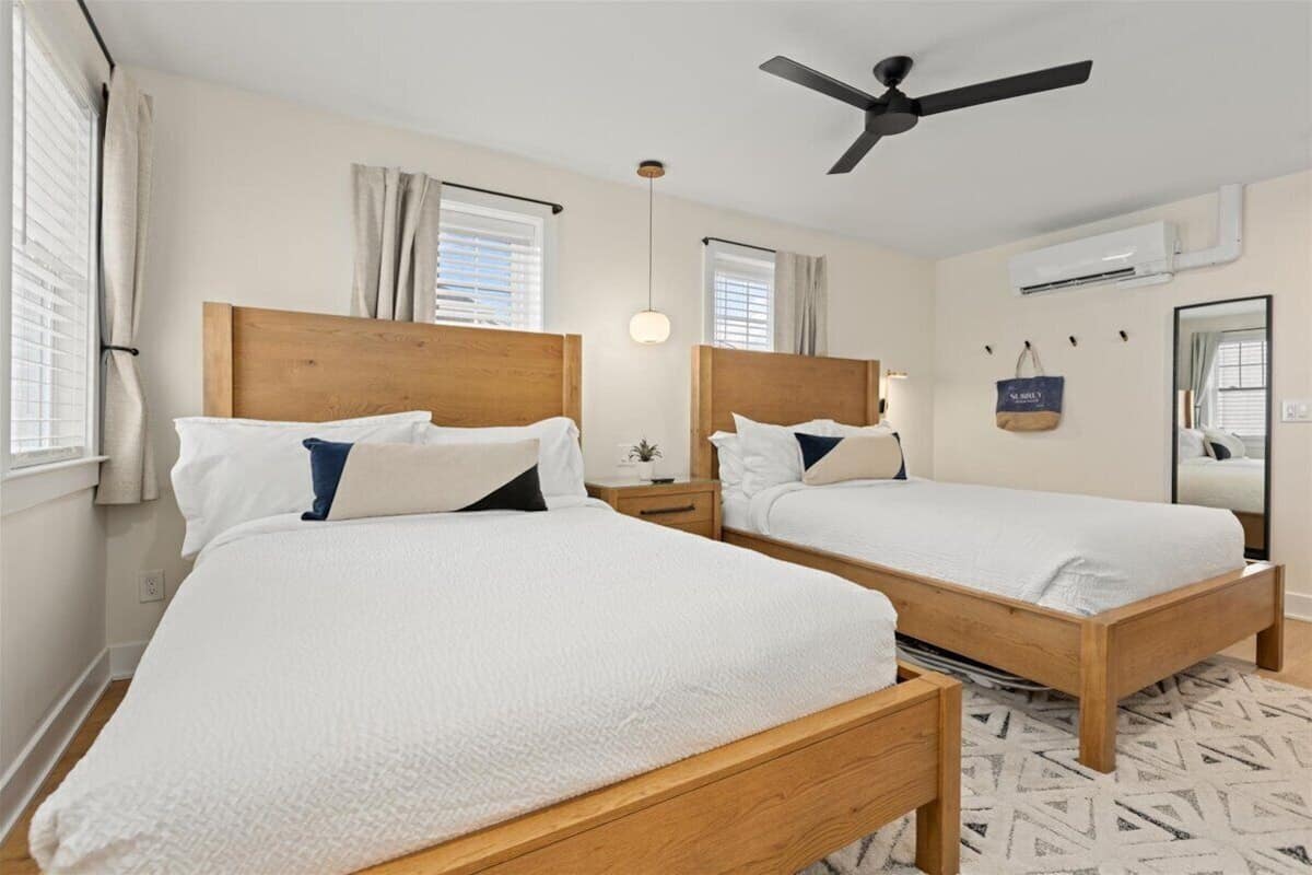 A hotel room with two wooden beds, a ceiling fan, an air conditioning unit, a bedside table with a lamp, and a patterned rug.