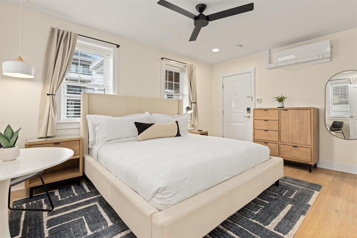 Une chambre à coucher comprenant un lit, des tables de chevet, un bureau, un miroir, une armoire et un ventilateur de plafond.
