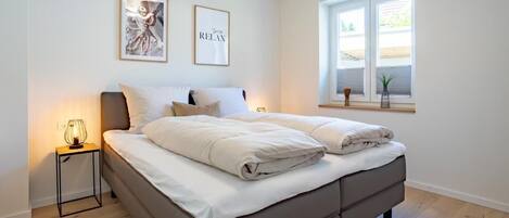 A bedroom with a bed, a bedside table with a lamp, two framed pictures on the wall, and a window with a view.