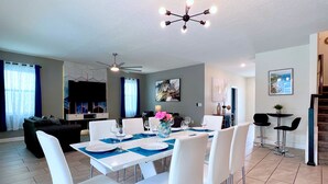 A modern dining area with a white table set for six, surrounded by white chairs. There is a ceiling fan above the table, and a TV is mounted on the wall to the left. - Avengers Game Room • Themed Rooms • South-Facing Pool & Spa • Near Disney (Kissimmee)