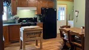 A kitchen with wooden cabinets, a black refrigerator, a wooden table, and a dining area with chairs. - Woof pack shack #1 (West Leyden)