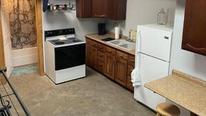A compact kitchen with wooden cabinets, a white stove, a sink, and a refrigerator. There is a small wooden table with stools. - Woofpackshack #4 (West Leyden)