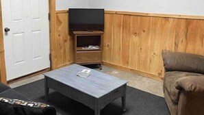 A room with a wooden paneled wall, a white door, a flat-screen TV on a wooden stand, a grey sofa, and a grey coffee table. - Woofpackshack #4 (West Leyden)