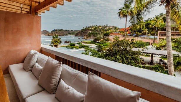 Un balcon avec un canapé et des coussins de couleur beige, donnant sur un paysage côtier avec des palmiers et une vue sur l’océan.