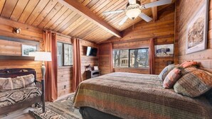 Un dormitorio acogedor de estilo cabaña con techo de madera, una cama con ropa de cama estampada, una mesita de noche, una lámpara y un televisor.