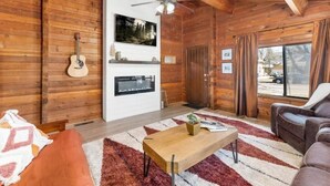 Interior acogedor de una cabaña con paredes de madera, chimenea, ventilador de techo y un gran ventanal.