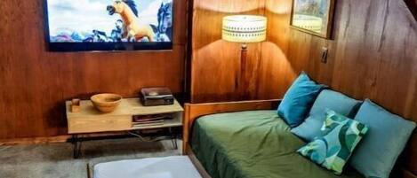 Una habitación con una cama de madera, televisión, lámpara y una mesita con un bol y un control remoto.