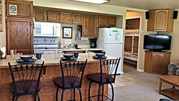 A compact kitchen with wooden cabinets, a central island with chairs, a refrigerator, and a television.