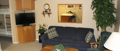 Ein Wohnzimmer mit einem dunkelblauen Sofa, einem Holztisch und einem Fernseher. Eine große Pflanze, eine Holzkommode und ein Spiegel sind an der Wand angebracht.