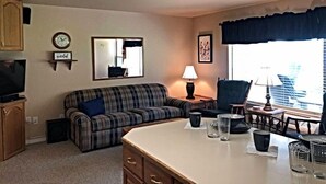 A living room with a plaid sofa, a small table with glasses, a lamp, and a window with a view of buildings.