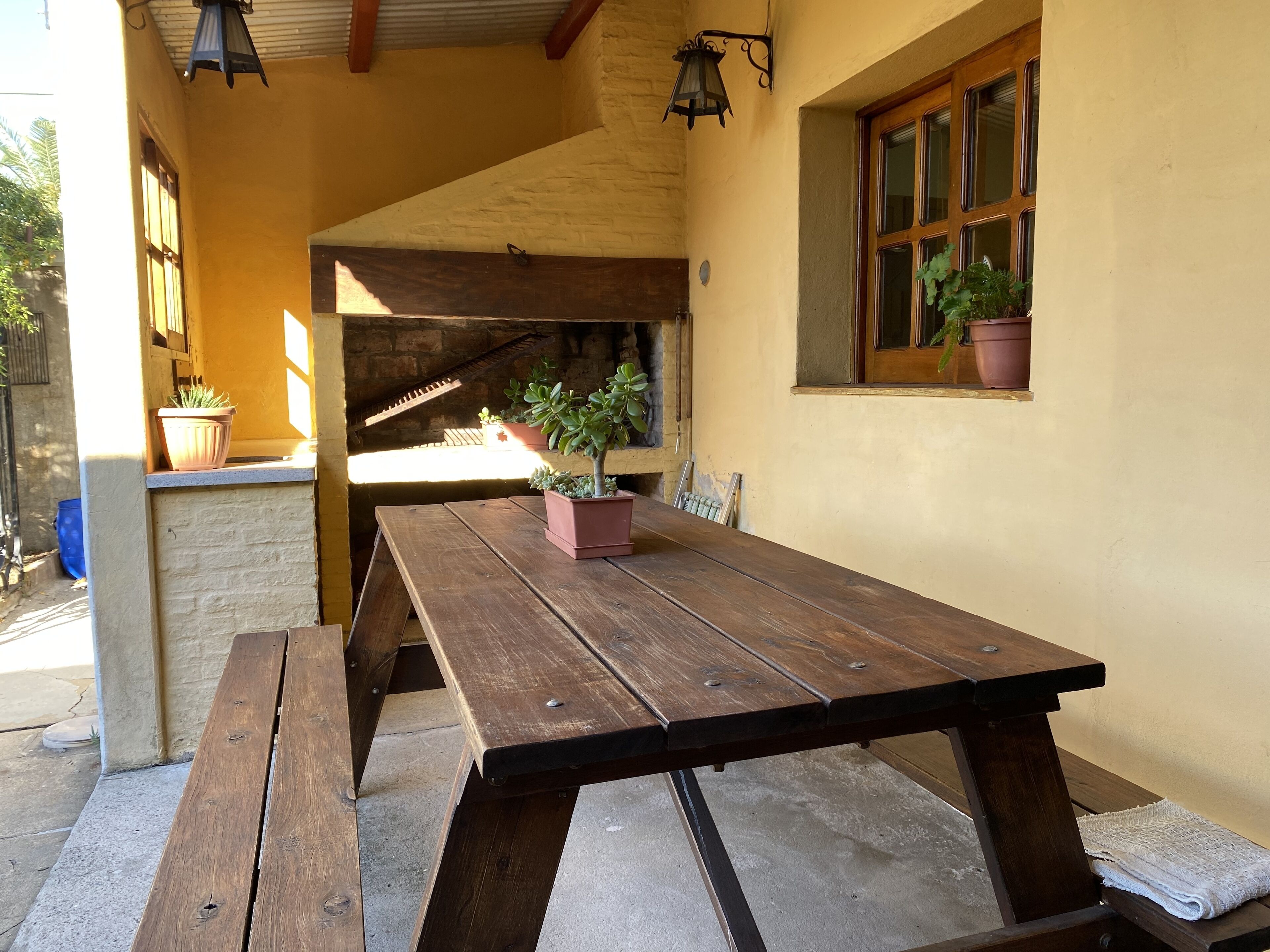 Une table en bois avec des bancs, installée dans un espace extérieur agrémenté d’un mur en pierre et d’une fenêtre ornée de plantes en pot.