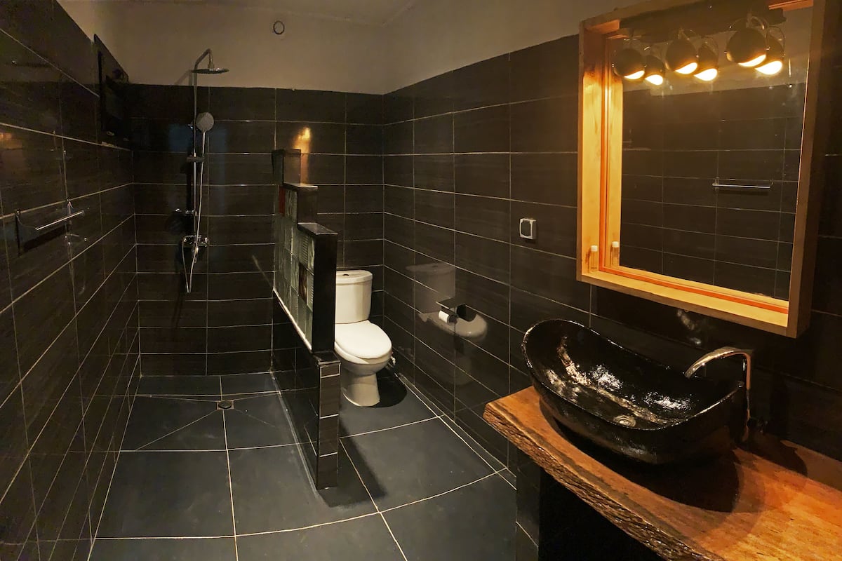 Un baño moderno con azulejos negros, un inodoro blanco, un lavamanos negro y un espejo con iluminación.