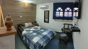 A bedroom with a bed, a nightstand, a lamp, a mirror, a window with arched design, an air conditioner, and a small stool.