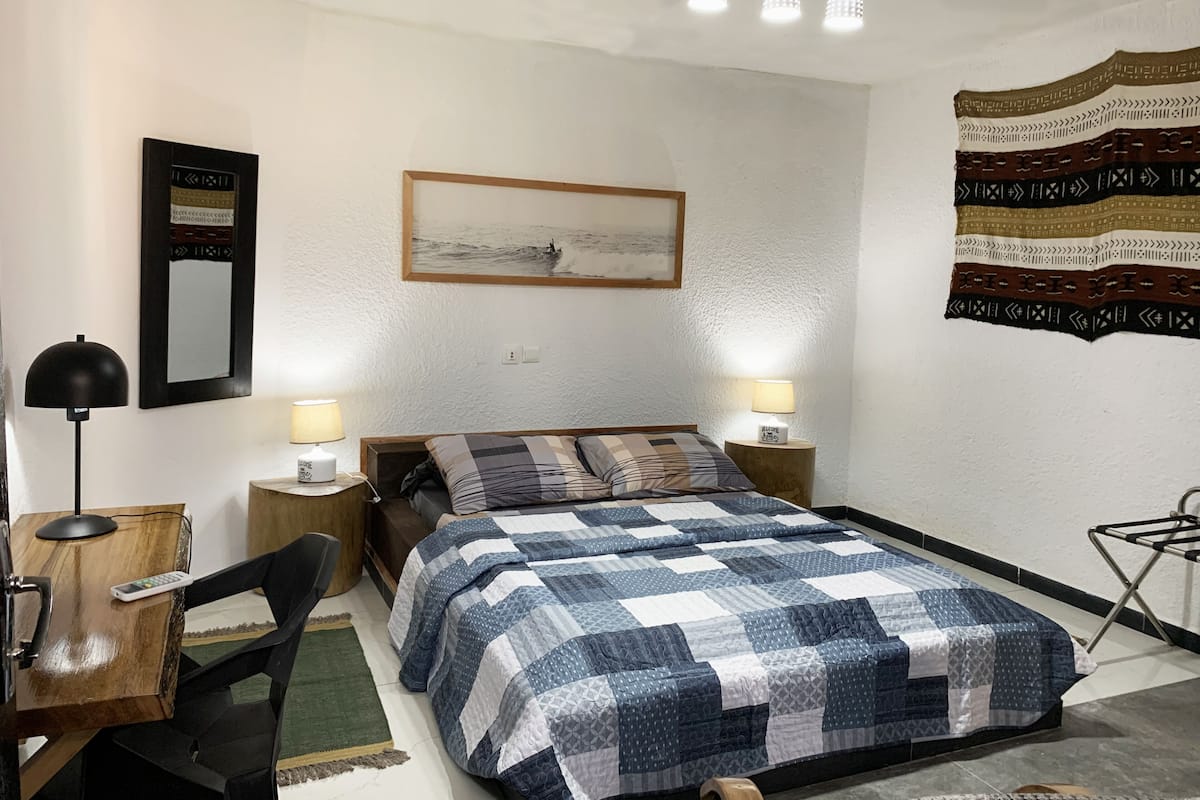 Un dormitorio con cama, dos lámparas de noche, un escritorio con lámpara, una silla, un tapete, un adorno en la pared y un cuadro enmarcado.