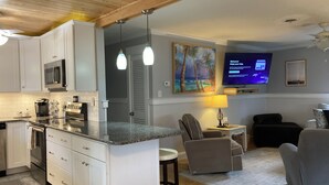 A modern kitchen with white cabinets, a granite countertop, and stainless steel appliances. There is a dining area with a gray chair and a small table. A flat-screen TV is mounted on the wall, displaying a welcome message. - Pet-Friendly OBX Retreat - 3BR/2BA -  Private Beach Parking MP6 Kill Devil Hills (Kill Devil Hills)