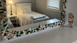 A bedroom with a bed, a nightstand, a lamp, a ceiling fan, a window with blinds, and a mirror with a seashell and starfish border. - Pet-Friendly OBX Retreat - 3BR/2BA -  Private Beach Parking MP6 Kill Devil Hills (Kill Devil Hills)