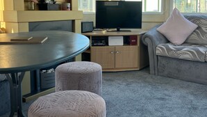 Een woonkamer met een ronde tafel, poefs, een bank met gestreepte kussens, een televisie en een kast met elektronische apparaten.