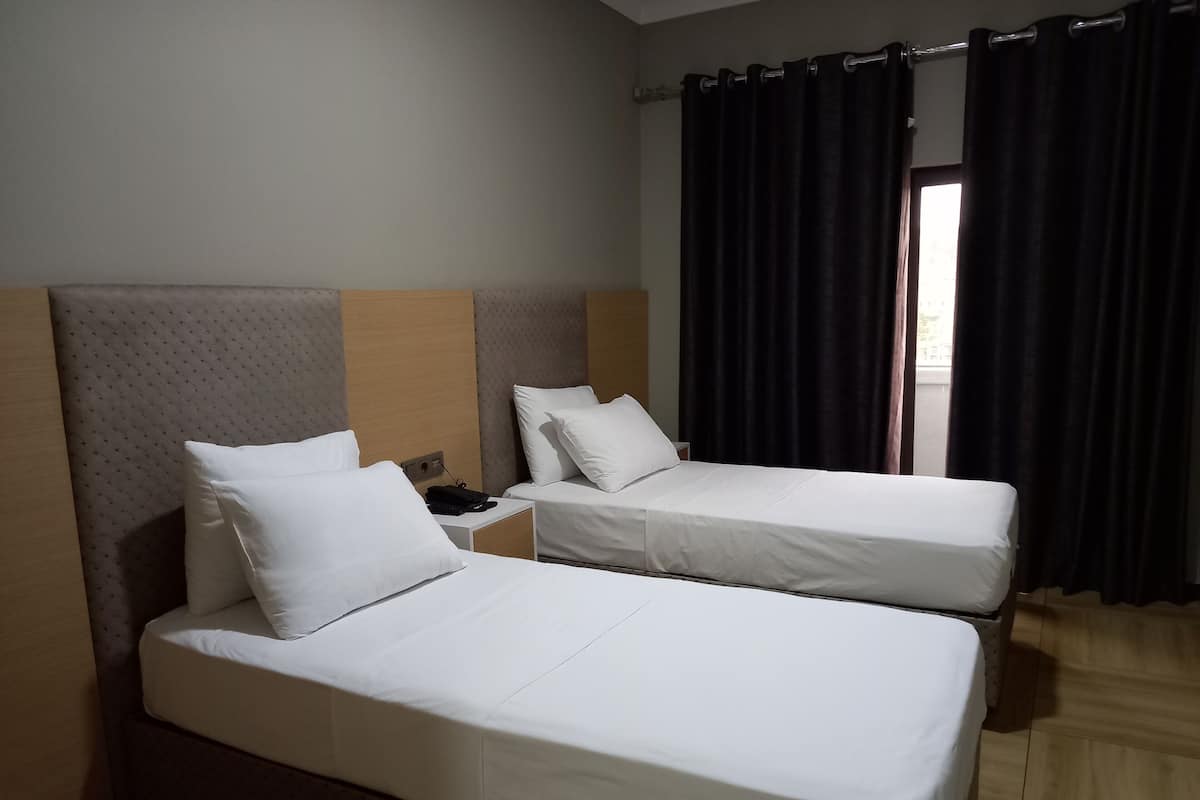 A hotel room with two single beds, a wooden headboard, a nightstand with a phone, and a window with dark curtains.
