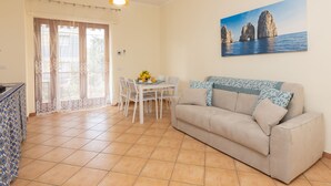 Ein Wohnzimmer mit einem beigefarbenen Sofa, einem Esstisch mit Stühlen, einem Gemälde von Küstenfelsen und einem Fenster mit Vorhängen.