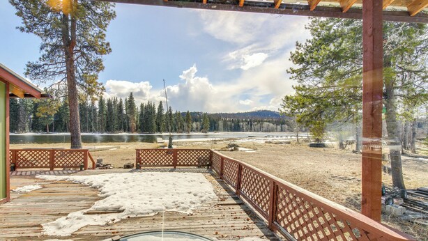 A wooden deck with a glass balustrade overlooking a lake, surrounded by trees and a clear sky. - Hike, Bike & Boat: Cabin w/ Dock on Seeley Lake! (Seeley Lake)