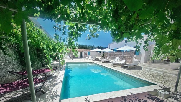 Ein Swimmingpool mit Liegestühlen und Sonnenschirmen, umgeben von Grünpflanzen und einem Steinweg.
