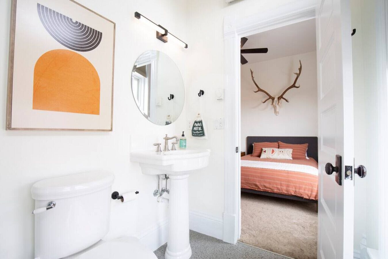 Baño con inodoro, lavabo y espejo. Se ve una cama a través de una puerta abierta, decorada con una cabeza de venado.
