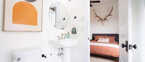 Baño con inodoro, lavabo y espejo. Se ve una cama a través de una puerta abierta, decorada con una cabeza de venado.